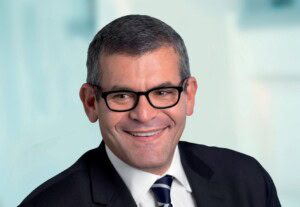 Brad-Fried-Picture.jpg Sir Bradley Fried, a man with short dark hair and glasses, is smiling while wearing a suit and striped tie against a blurred light background.