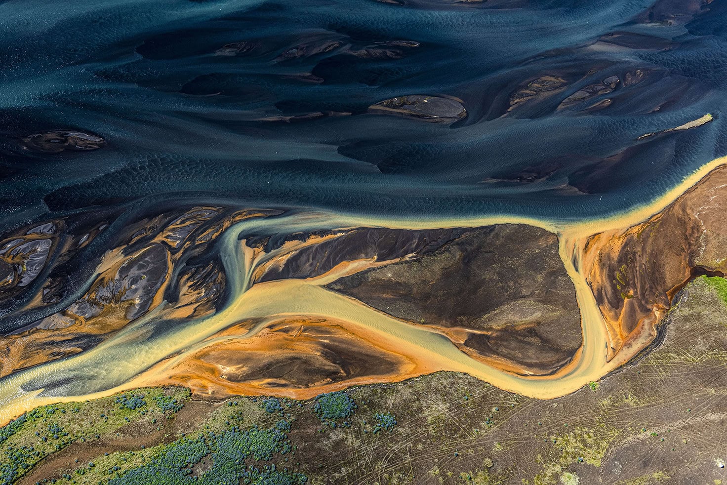 Aerial view of a river delta with winding, multicolored sediment paths meeting dark water, reminiscent of intricate networks designed for institutional clients, and a patch of green vegetation.