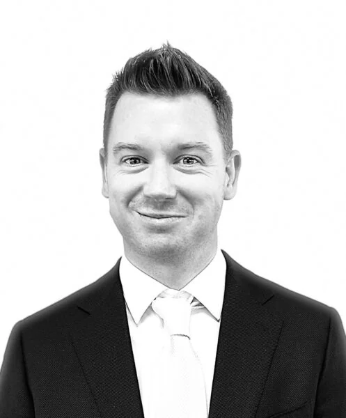 Black and white portrait of Robert Skelton, a smiling man in a suit and tie, with short hair combed upwards.