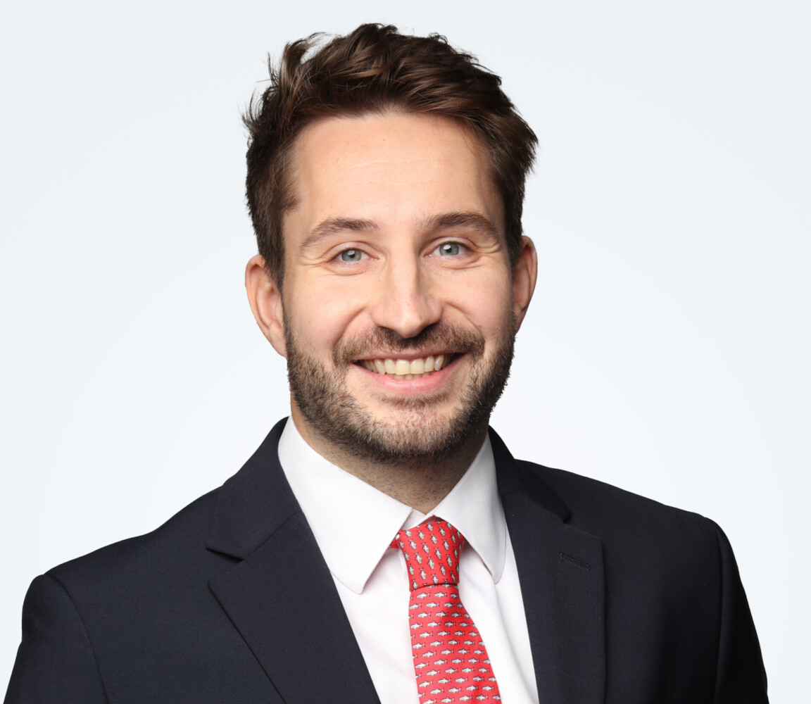 Johnny Meldram, in a dark suit and red tie, smiles confidently at the camera against a plain white background.