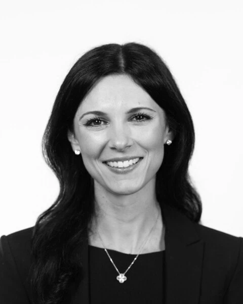 A black and white image captures Pamela Haddad, a woman with long dark hair, wearing a suit jacket and necklace, smiling against a plain background.