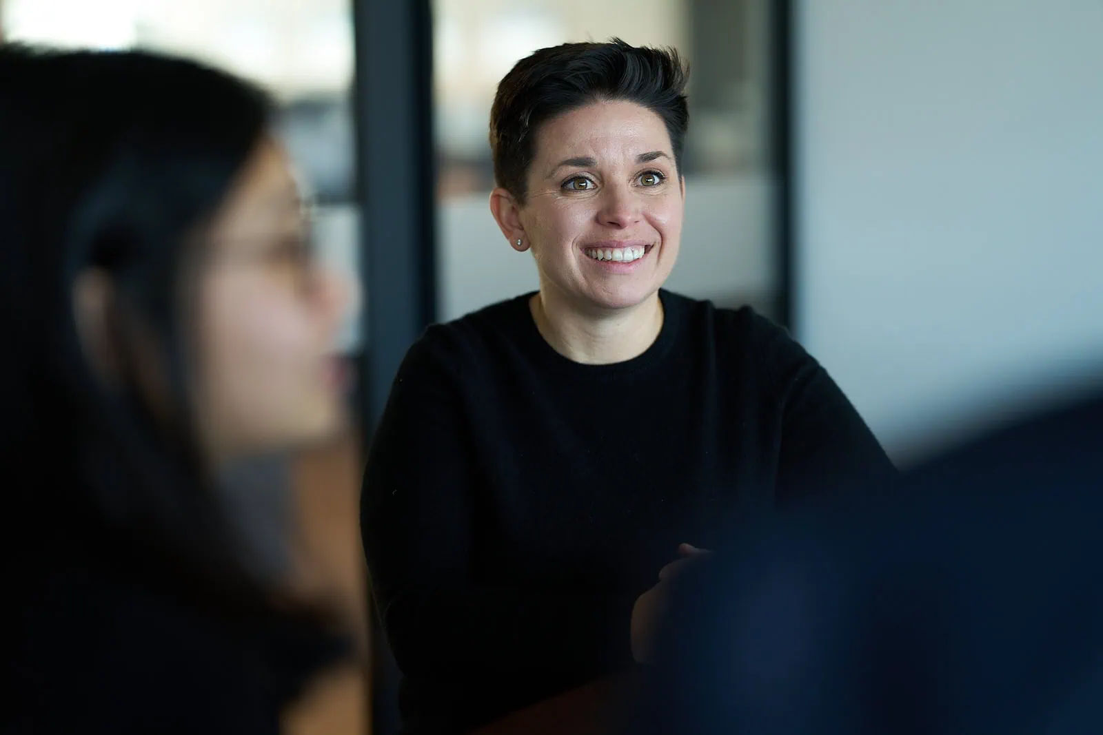 A person with short hair, wearing a black shirt, smiles while sitting at a table in a meeting room focused on promoting diversity and inclusion. Blurred individuals are in the foreground.