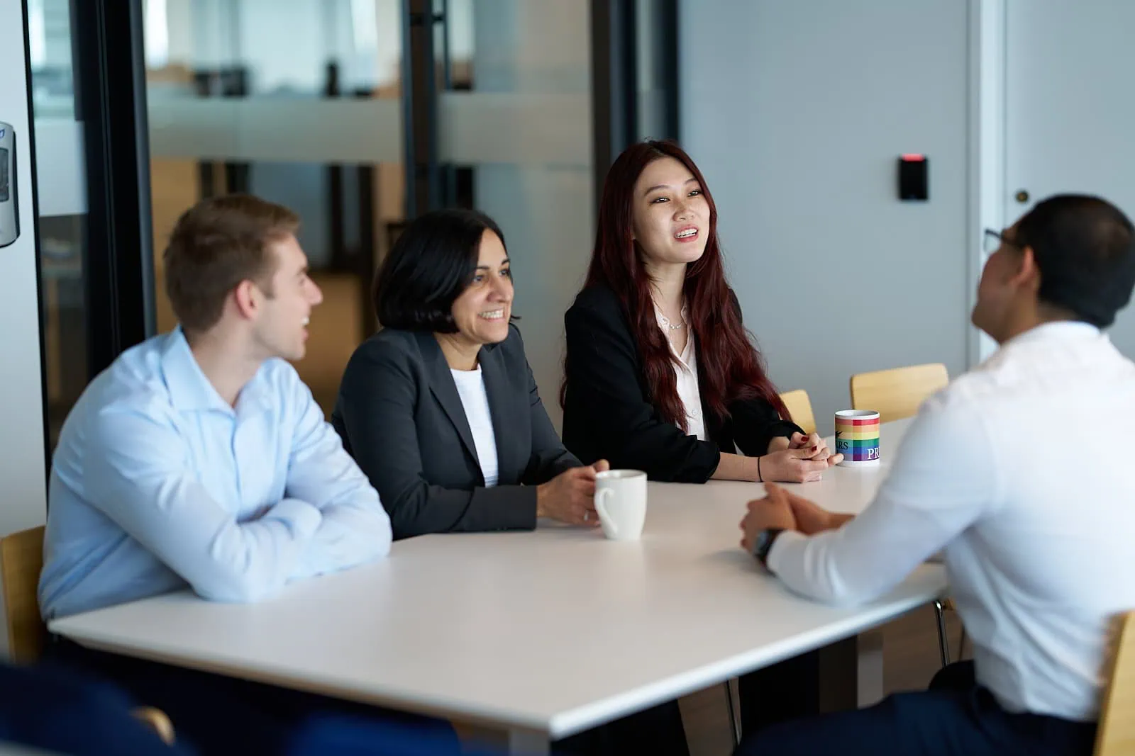 Four partners gather around a table in a Boston office setting, engaged in lively conversation. Two hold mugs, emphasizing the comfortable yet professional atmosphere of this capital-driven meeting.