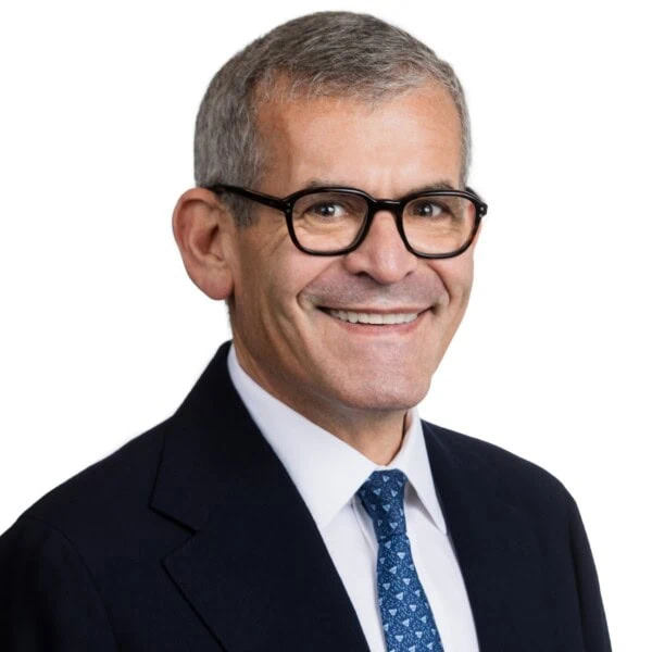 Sir Bradley Fried, a man with short gray hair, wearing glasses, a dark suit jacket, white shirt, and patterned blue tie, smiles against a plain white background.