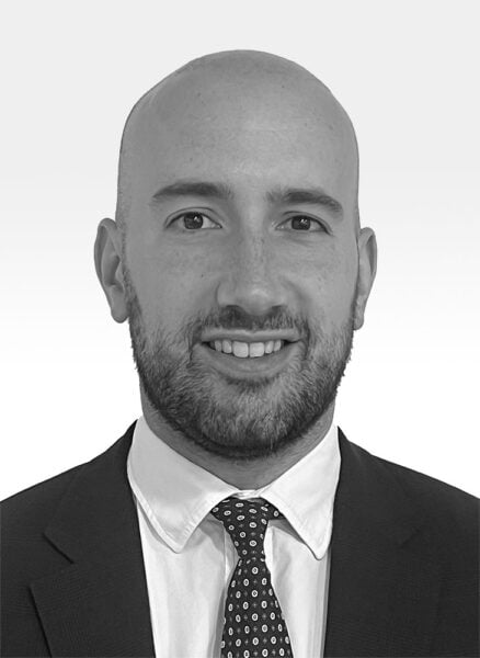 Joe Basrawy, a bald man with a beard, is wearing a dark suit, white shirt, and patterned tie. He is smiling slightly and looking directly at the camera against a plain background.