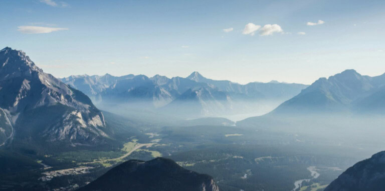 An aerial view of a mountain range with valleys and peaks under a clear blue sky, where mist envelops the lower areas, reminds us of the untouched beauty we aim to preserve through sustainable investing.