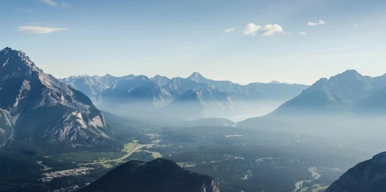 Mountain range with peaks and valleys under a clear sky, partially covered in mist, with forests and open areas visible below—a serene landscape that echoes the harmony sought in Sustainable Investing for 2024-2025.