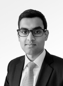 A black and white portrait of Amar Patel with short hair, wearing glasses, a suit, and a patterned tie, looking directly at the camera.