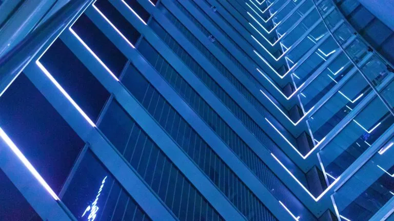 Close-up view of a tall modern building at night, with blue-tinted glass panels and illuminated lines highlighting the structure's geometric design—a primer in urban architecture.