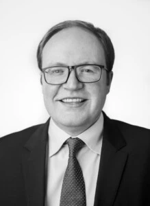 Euan Finlay, wearing glasses, a suit, and a tie, smiles against a plain white background in a black and white photograph.