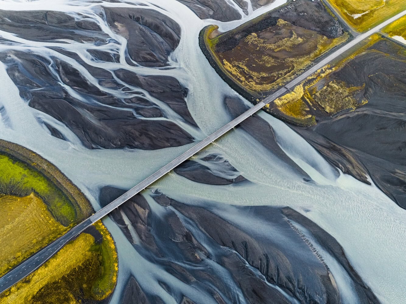 Aerial view of a long bridge crossing a river with multiple branches, surrounded by dark volcanic terrain and patches of green moss—an inspiring landscape inviting insights for 2025.