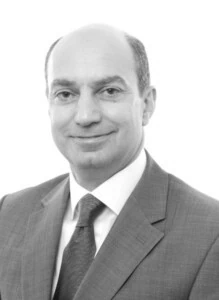 A bald man in a suit and tie named Kamran Moghadam, smiling at the camera against a white background,