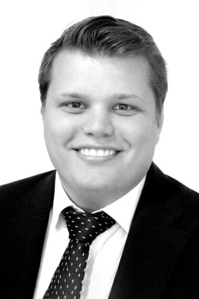 A black and white portrait of Matt Libling. He is a man with short hair, smiles while wearing a dark suit, white shirt, and polka-dotted tie, set against a plain background.