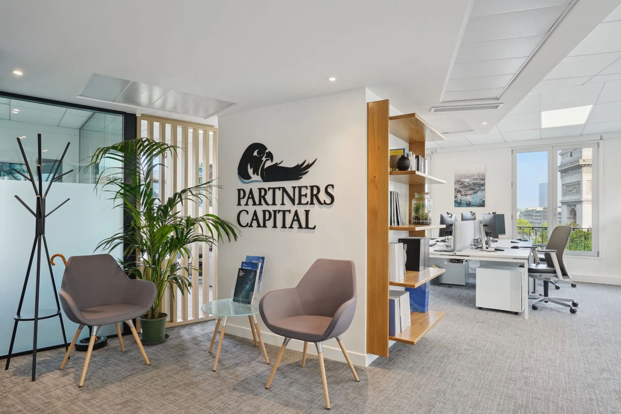 Modern office space with the "Partners Capital Paris" logo on the wall, two chairs around a small table with magazines, a coat rack, a potted plant, and desks with computers in the background.