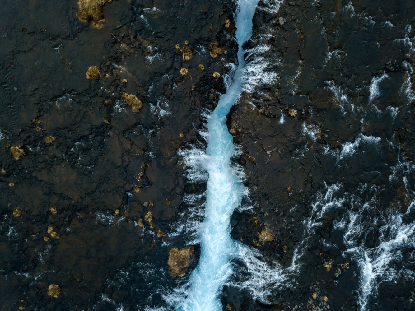 An OCIO aerial view reveals a narrow river meandering through dark, rocky terrain, highlighting the stark contrast between the bright blue water and the surrounding landscape.