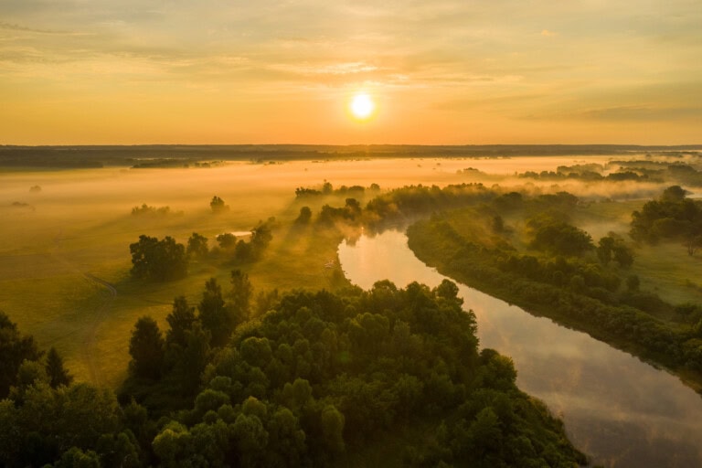 Aerial view of a winding river surrounded by lush greenery at sunrise, with mist floating over the landscape—a scene that embodies the spirit of sustainable investing in 2024-2025.