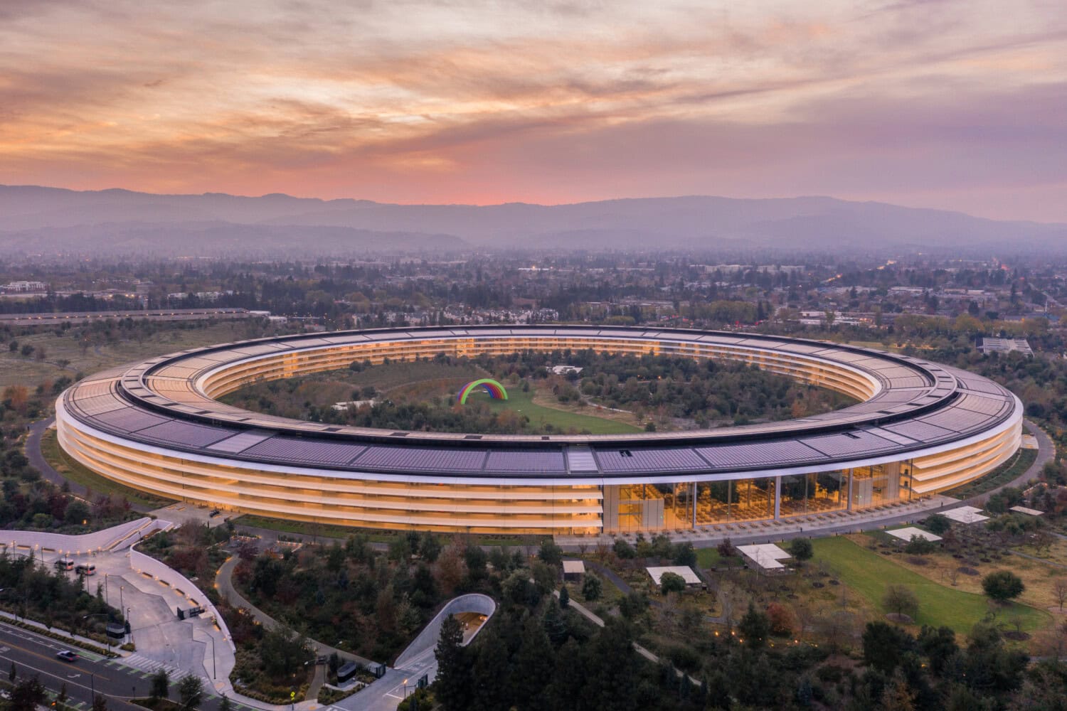 Aerial view of a circular, futuristic building nestled in landscaped grounds, surrounded by roads and trees under a cloudy sky at sunset—a visionary hub for tech entrepreneurs seeking inspiration.