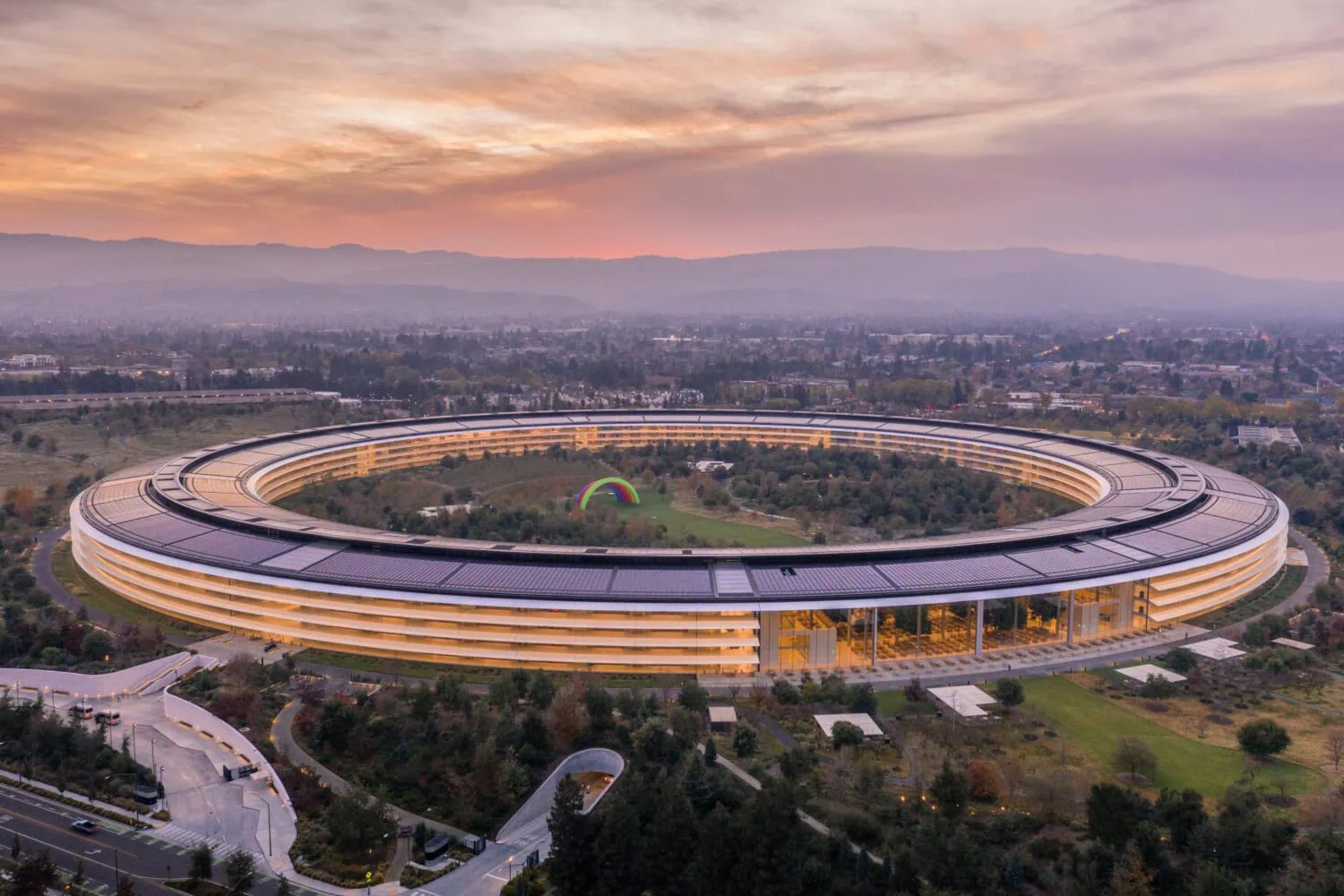 Aerial view of a circular, futuristic building nestled in landscaped grounds, surrounded by roads and trees under a cloudy sky at sunset—a visionary hub for tech entrepreneurs seeking inspiration.