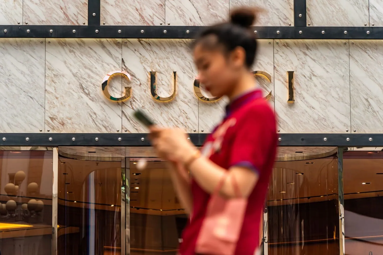 A blurred person walks by a Gucci store, phone in hand, as the name glistens in the background—a subtle nod to the luxury market's resilience amid whispers of a China stimulus.