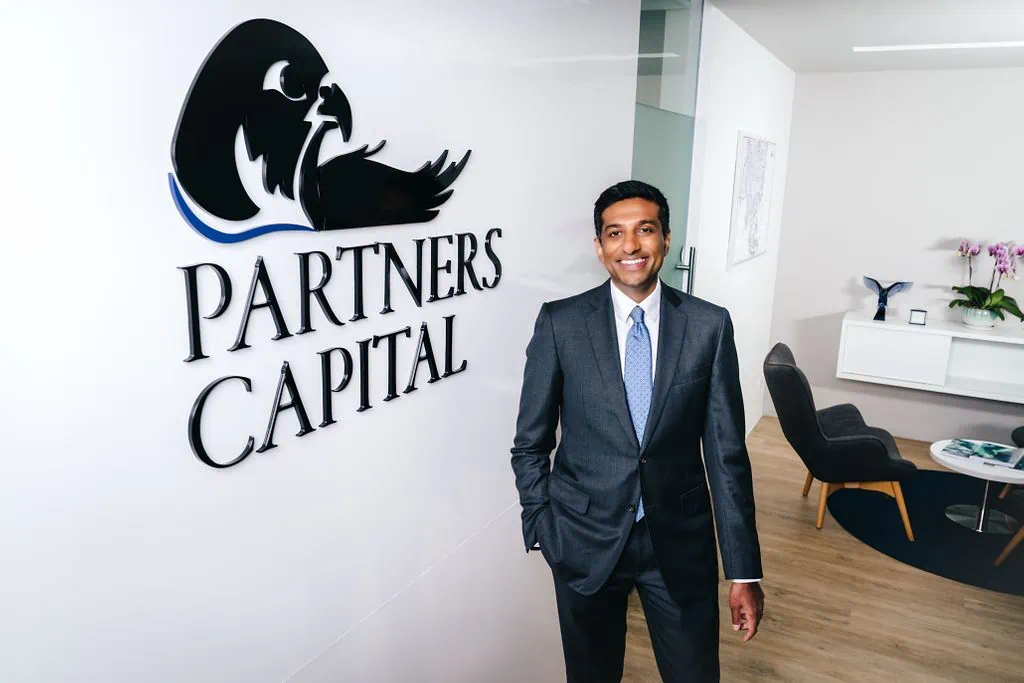 A man in a suit stands smiling in an office lobby with an "OCIO Partners Capital" logo on the wall behind him.